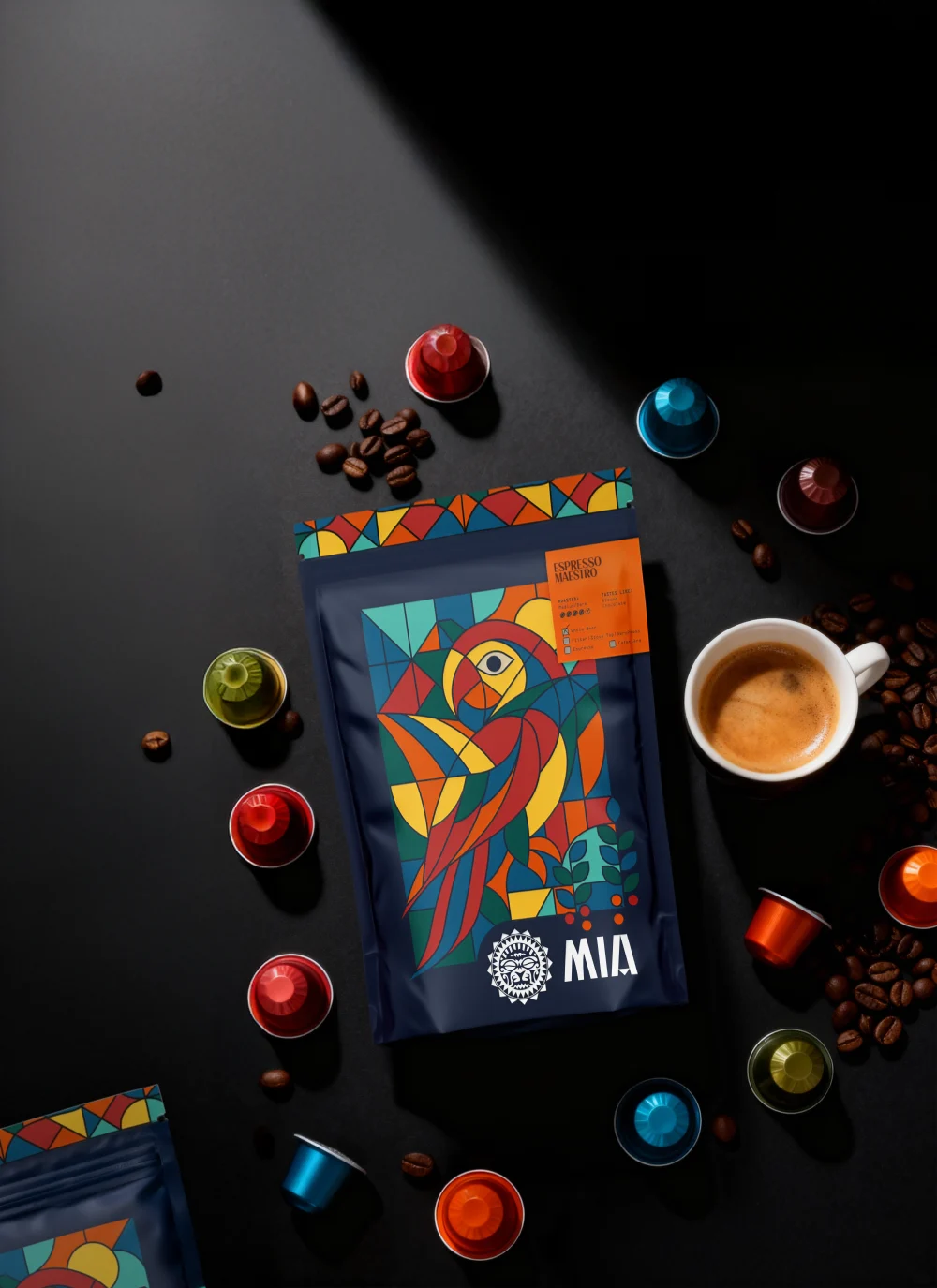 Dark flat-lay of a Mia parrot-illustrated coffee bag surrounded by espresso pods, beans, and a fresh espresso